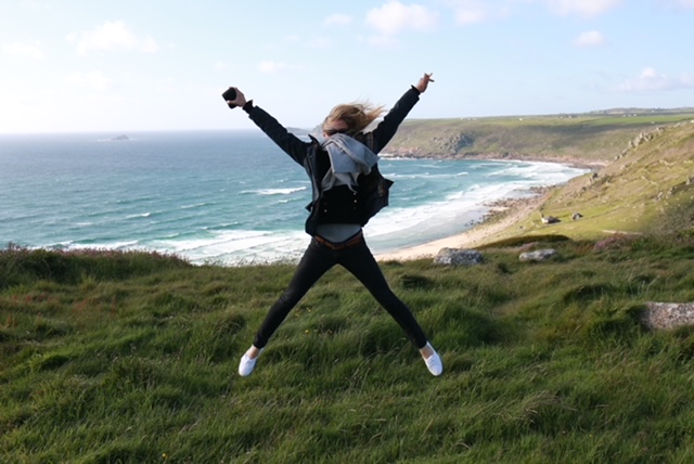 Gemma Jennings jumps for joy at the sight of the sea. She is standing on a rugged coastline, with rocky scree down to the beach below.