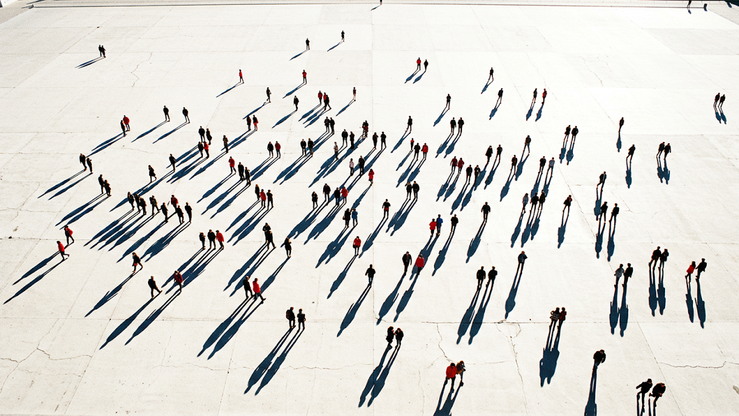 High-angle aerial shot of a scattered crowd walking across a vast, white concrete plaza, casting long, sharp shadows due to bright, low-angle sunlight.