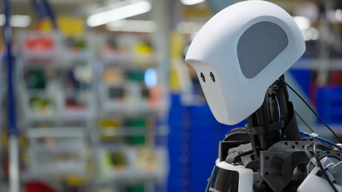 a close-up of the smooth, white head and exposed mechanics of a humanoid robot looking to the left, with a blurred backdrop of an industrial or warehouse setting with shelves and storage bins.