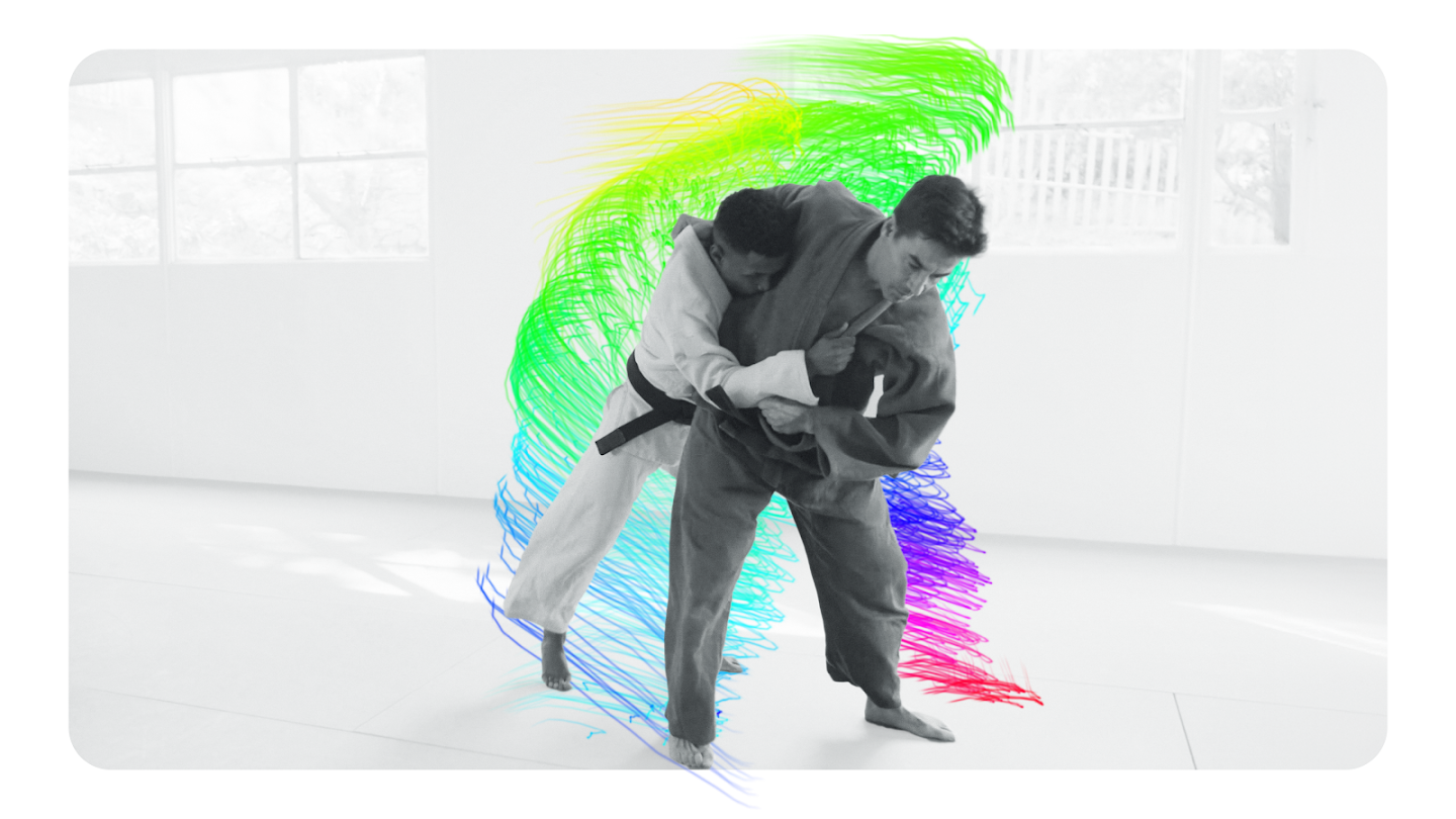 An image showing two people in a martial arts studio (dojo) practicing judo or jiu-jitsu. They are mid-motion in a grappling hold, surrounded by a vibrant, rainbow-colored light trail that follows their movement path. The background is a clean, bright white studio with windows.