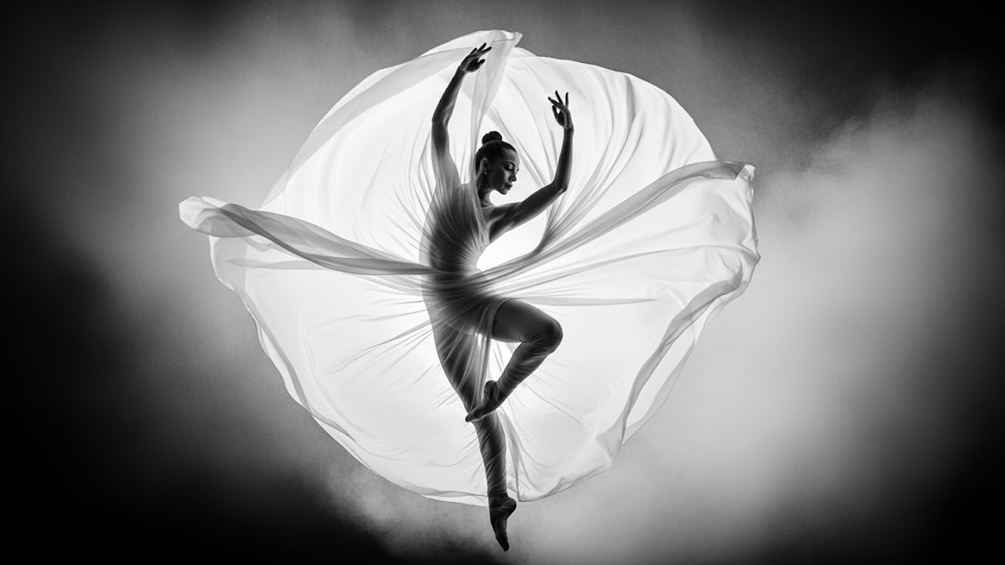 AI-generated image of: A dramatic black and white photograph of a dancer silhouetted against bright backlighting, mid-leap. Large, flowing sheer fabric billows around the dancer, creating a circular, ethereal effect.